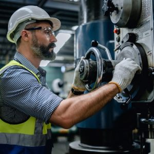 An engineer wearing a safety vest and hard hat turns a knob on a machine in the middle of a warehouse.