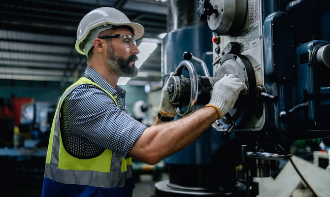 An engineer wearing a safety vest and hard hat turns a knob on a machine in the middle of a warehouse.