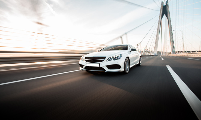 A sleek white sedan drives alone over a highway bridge during the day. The surroundings are blurred in motion.