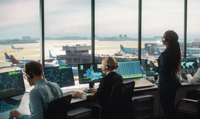 Four air traffic controllers monitoring computer screens in a control tower with curved glass walls.