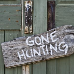 A weathered sign reading ‘Gone Hunting’ written in white paint hangs outside a light green rustic building.