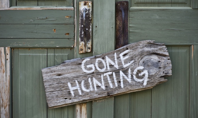 A weathered sign reading ‘Gone Hunting’ written in white paint hangs outside a light green rustic building.