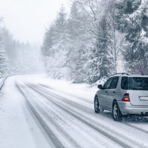 A silver SUV drives along a snow-covered road lined with a guardrail and surrounded by tall, mature trees.
