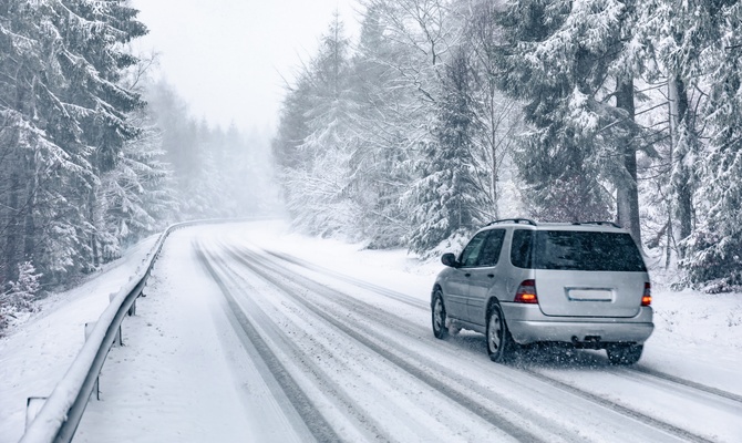 A silver SUV drives along a snow-covered road lined with a guardrail and surrounded by tall, mature trees.