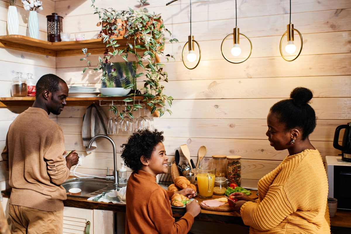 A man, a woman, and a young boy cook together in a rustic kitchen featuring shiplap walls and wooden shelves.