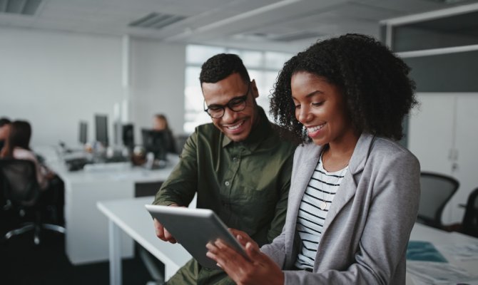 Two young Black business professionals are smiling as they work together in a shared office computer space.