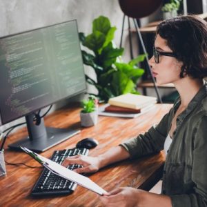 A young professional sits at her workspace with a dual monitor setup and a headset on. She's holding a clipboard.