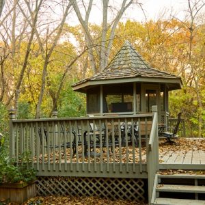 A backyard deck and matching gazebo built off a home, surrounded by a forest with leaves changing to autumn colors.