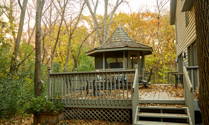 A backyard deck and matching gazebo built off a home, surrounded by a forest with leaves changing to autumn colors.