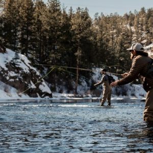 A pair of anglers in outdoor gear casting flies for fly fishing in a chilly Colorado river surrounded by trees and snow.