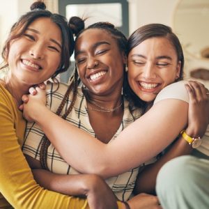 Three smiling young women sitting on a tan living room couch with their arms wrapped around each other.