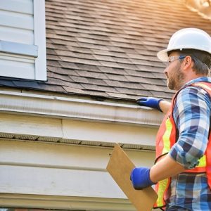 A man wearing a white hard hat and safety vests holds a clipboard and inspects the black roof of a house.