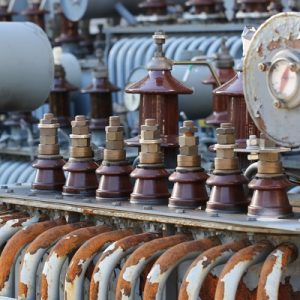 A close-up of a power transformer in a power plant. The silver pipes have numerous patches of reddish-brown rust.