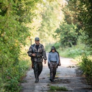 A hunter walks with his younger child through a rural area. Both are wearing hunting clothes and shoes.