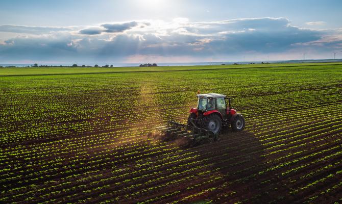 An aerial view of a red tractor cultivating a large field of green crops as the sun shines through the clouds.
