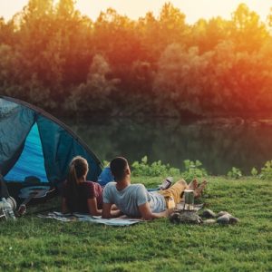 A Man and a woman lying on top of lush grass next to a large blue tent overlooking a river and multiple trees.