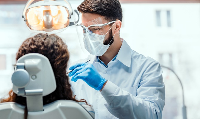 A man wearing a white lab coat and blue rubber gloves holding a tool near a woman's mouth as she sits in a dentist's chair.
