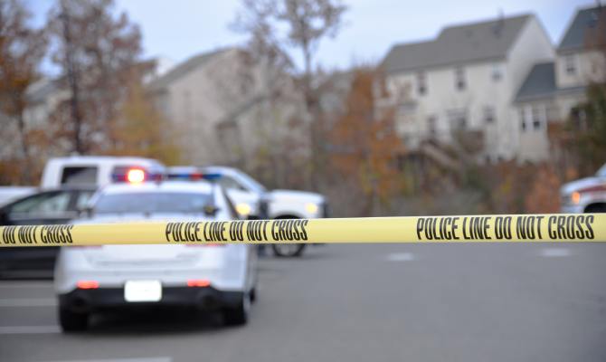 A cop car in a crowded parking lot. A yellow piece of tape surrounds the lot and reads “Police Line Do Not Cross.”