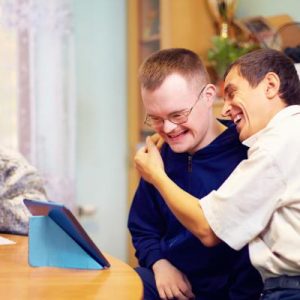 Two people sitting at a table and hugging while looking at a tablet. Another person writes at the table.