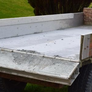 An old, beat up hauling trailer sits parked in a grassy field. The trailer's tailgate is open and dirty with mud.