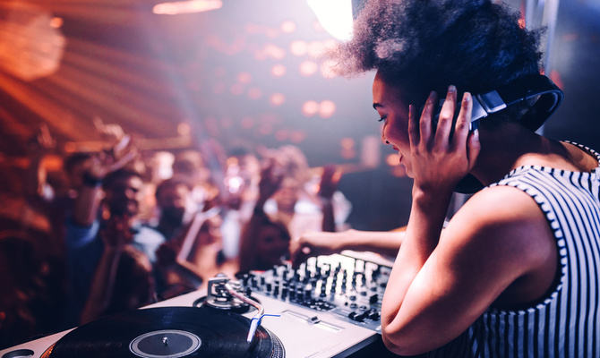 A female DJ in a striped tank top wears headphones as she uses her equipment to play music for an energetic crowd.
