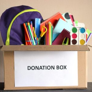 A cardboard box full of school supplies with a white sheet of paper in the front, displaying the words “donation box”.