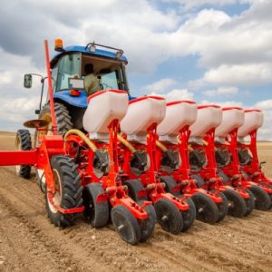 A blue tractor is using a red seed drill attachment to sow crop seeds in a tilled field.
