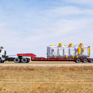 A side view of white semi-truck with a flatbed hauling a large rectangular industrial container on an empty road.