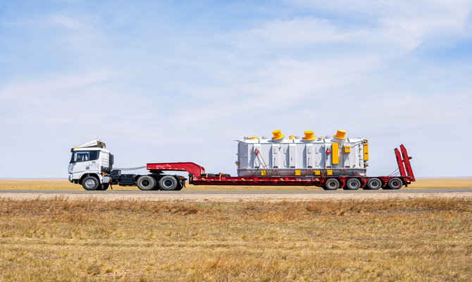 A side view of white semi-truck with a flatbed hauling a large rectangular industrial container on an empty road.