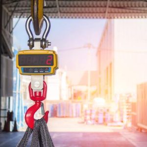 A close-up of a yellow industrial crane scale with a red hook lifting the straps of a load inside a warehouse.