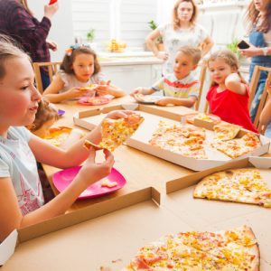 A young girl is eating a slice of pizza at a table surrounded by other kids with adults in the background.