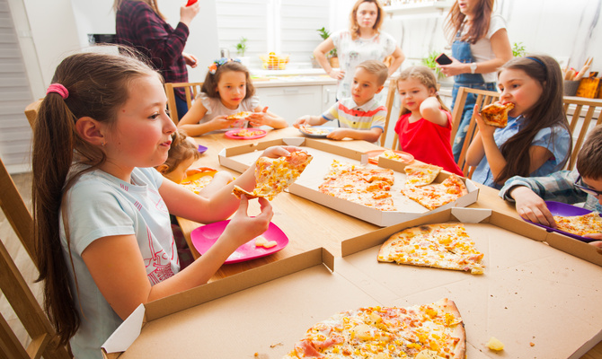 A young girl is eating a slice of pizza at a table surrounded by other kids with adults in the background.