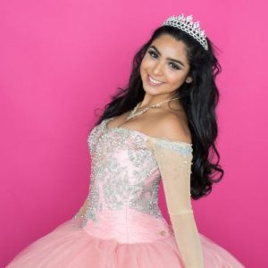A young woman wearing a formal dress and tiara stands against a solid background while facing forward with a slight turn.