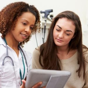 A teenage girl wearing a tan sweater is looking at a tablet that a female doctor is holding in a healthcare room.