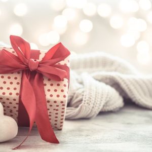 A red and white polka dot giftbox wrapped with a red ribbon sits next to a white heart and a knitted blanket.