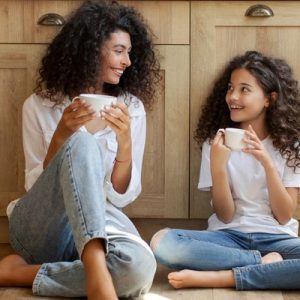 A mother and daughter sit on the kitchen floor in front of some cabinets. They both hold a white coffee cup.
