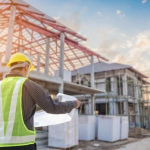 Construction worker in a hard hat holding blueprints at a house building site with framing visible in the background.
