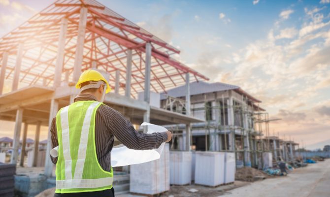 Construction worker in a hard hat holding blueprints at a house building site with framing visible in the background.
