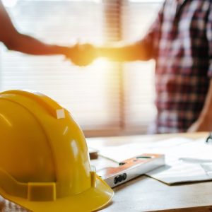 A yellow safety helmet rests on a desk with papers and tools while two people shake hands in the background