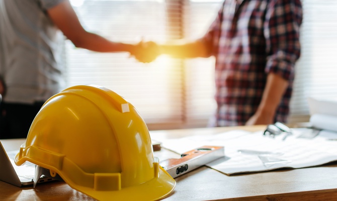A yellow safety helmet rests on a desk with papers and tools while two people shake hands in the background