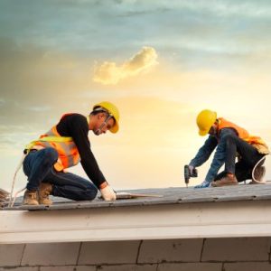 Two construction workers in safety gear use tools to install metal roofing panels on a sloped roof at sunset.