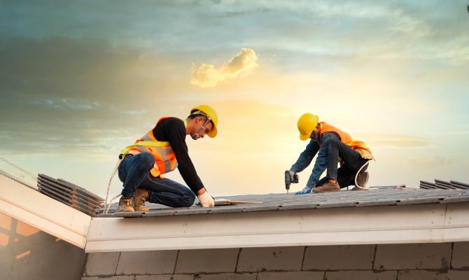 Two construction workers in safety gear use tools to install metal roofing panels on a sloped roof at sunset.