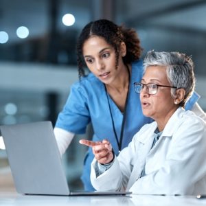 A doctor sitting and pointing at a laptop on their desk. A nurse leans over their shoulder to look at the laptop screen.
