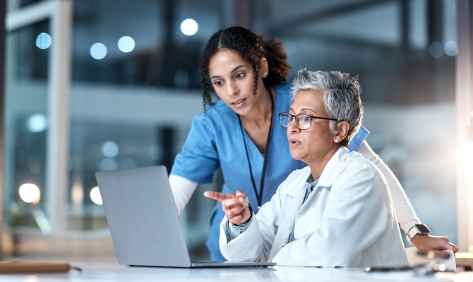 A doctor sitting and pointing at a laptop on their desk. A nurse leans over their shoulder to look at the laptop screen.