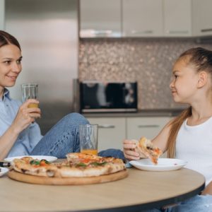 A mother and daughter sit at a kitchen table eating fresh pizza and talking while drinking juice together.