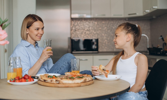 A mother and daughter sit at a kitchen table eating fresh pizza and talking while drinking juice together.