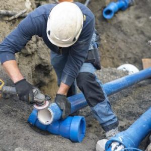 A group of workers installing underground pipes in a trench using equipment with exposed soil and utility lines visible.