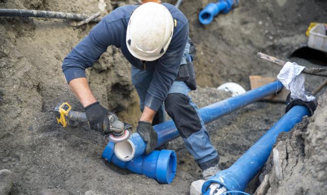 A group of workers installing underground pipes in a trench using equipment with exposed soil and utility lines visible.