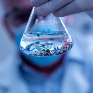 A gloved hand holding a flask with liquid and visible microplastic particles, with a scientist blurred in the background.