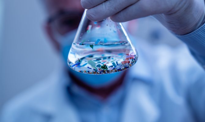 A gloved hand holding a flask with liquid and visible microplastic particles, with a scientist blurred in the background.